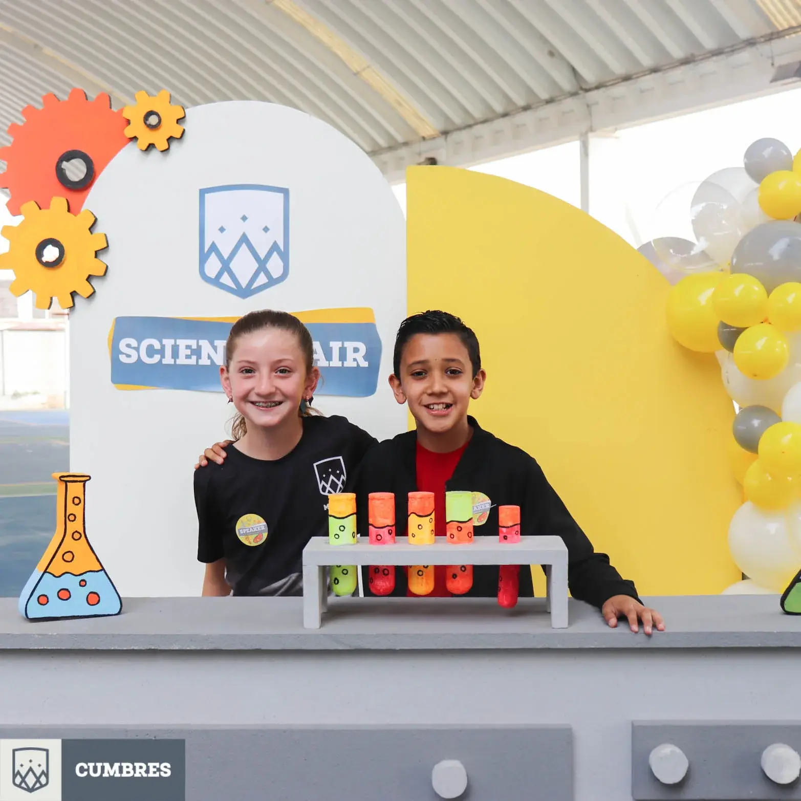 Dos estudiantes participan en una actividad de ciencias en la feria "Science Fair" con experimentos coloridos en la estación.